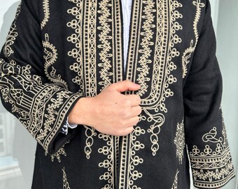 Può includere: Una giacca nera caratterizzata da intricati ricami color oro lungo l'apertura frontale, il colletto e le maniche. Il capo presenta dettagliati motivi geometrici e floreali sul tessuto, con una camicia bianca a colletto visibile sotto.