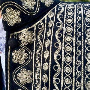 Pode incluir: Close-up de um casaco preto com intrincados bordados dourados. O design apresenta padr&otilde;es florais e bordas decorativas. Um bot&atilde;o preto &eacute; vis&iacute;vel na frente do casaco. A gola do casaco tamb&eacute;m &eacute; adornada com bordados dourados.