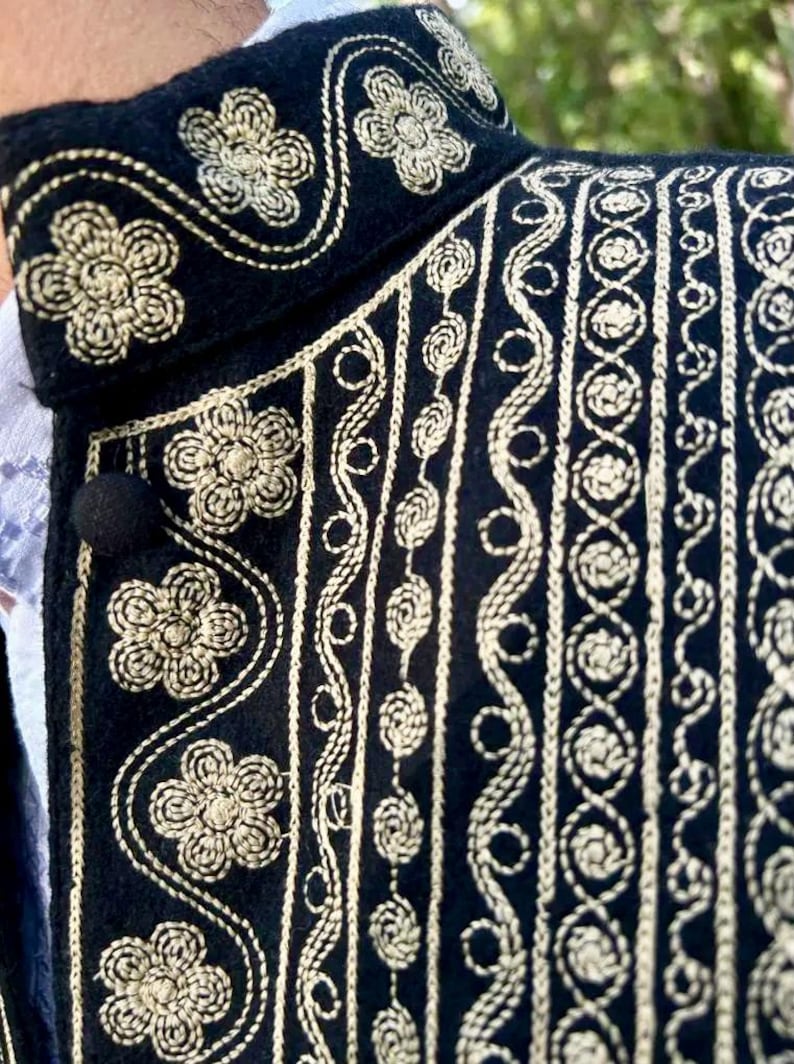 Pode incluir: Close-up de um casaco preto com intrincados bordados dourados. O design apresenta padr&otilde;es florais e bordas decorativas. Um bot&atilde;o preto &eacute; vis&iacute;vel na frente do casaco. A gola do casaco tamb&eacute;m &eacute; adornada com bordados dourados.