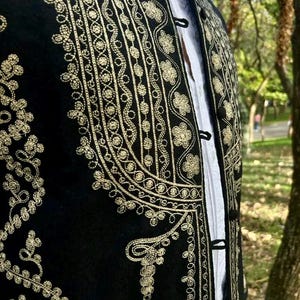 Pode incluir: Colete preto bordado com desenhos florais e de volutas douradas. O colete apresenta gola alta e fechos de bot&otilde;es. Uma camisa branca &eacute; vis&iacute;vel por baixo. A pe&ccedil;a de roupa faz provavelmente parte de um traje tradicional ou formal.