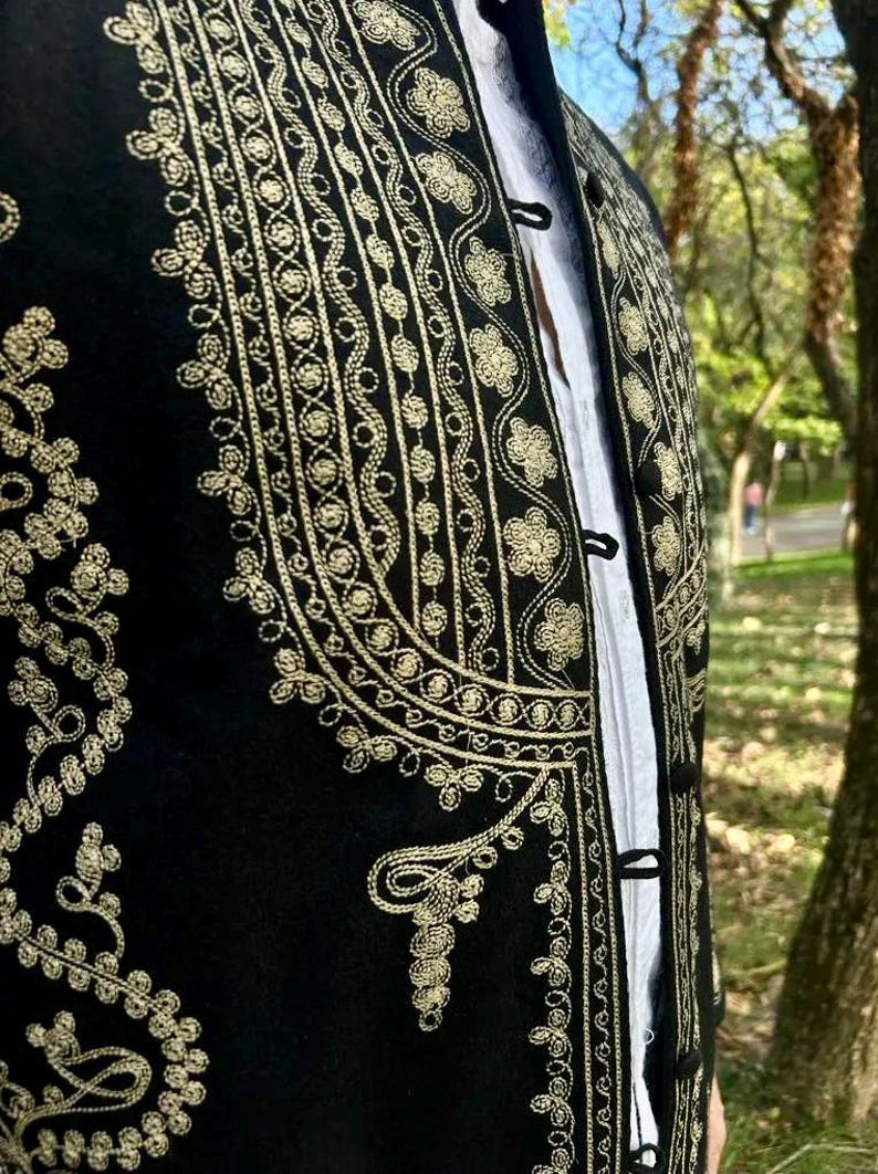 Pode incluir: Colete preto bordado com desenhos florais e de volutas douradas. O colete apresenta gola alta e fechos de bot&otilde;es. Uma camisa branca &eacute; vis&iacute;vel por baixo. A pe&ccedil;a de roupa faz provavelmente parte de um traje tradicional ou formal.