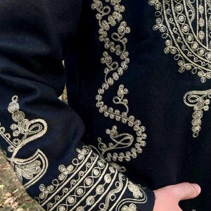 Pode incluir: Close-up de uma pe&ccedil;a de roupa preta com bordados dourados intrincados. O bordado apresenta padr&otilde;es florais e geom&eacute;tricos nas mangas e no corpete. O design &eacute; detalhado e ornamentado, sugerindo um estilo formal ou cerimonial.