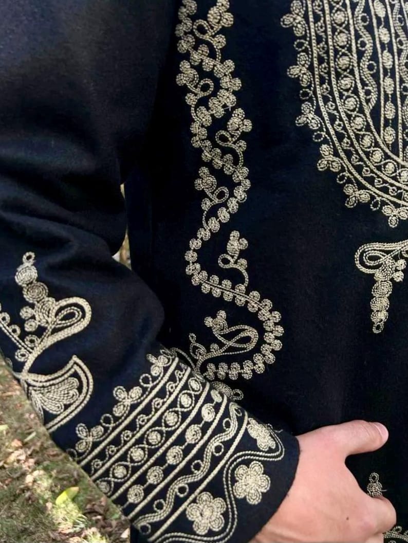 Pode incluir: Close-up de uma pe&ccedil;a de roupa preta com bordados dourados intrincados. O bordado apresenta padr&otilde;es florais e geom&eacute;tricos nas mangas e no corpete. O design &eacute; detalhado e ornamentado, sugerindo um estilo formal ou cerimonial.
