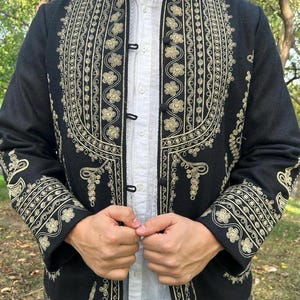 Pode incluir: Casaco preto com intrincados bordados dourados. O casaco apresenta padr&otilde;es florais e de rolagem ornamentados, com desenhos detalhados na gola, punhos e frente. Usado sobre uma camisa branca com gola.