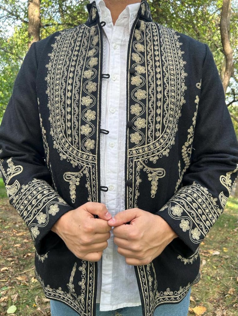 Pode incluir: Casaco preto com intrincados bordados dourados. O casaco apresenta padr&otilde;es florais e de rolagem ornamentados, com desenhos detalhados na gola, punhos e frente. Usado sobre uma camisa branca com gola.