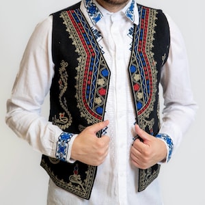 Könnte beinhalten: Eine schwarze Weste mit aufwendiger Stickerei in Rot, Blau und Gold. Sie wird über einem weißen Hemd mit passendem besticktem Kragen und Manschetten getragen. Das Design ist traditionell.