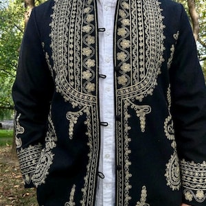 Pode incluir: Casaco preto com intrincados bordados dourados. O casaco apresenta padr&otilde;es florais e geom&eacute;tricos ornamentados na gola, na frente e nas mangas. &Eacute; usado sobre uma camisa branca e tem fechos de bot&otilde;es pretos. O design do casaco &eacute; detalhado e atraente.