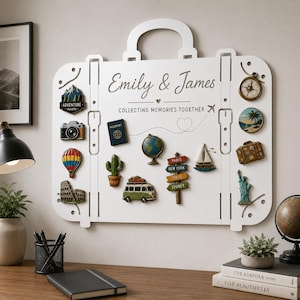 May include: A white suitcase-shaped wall display board featuring the text Emily & James and COLLECTING MEMORIES TOGETHER. The board is decorated with various travel-themed magnets including a camera, hot air balloon, globe, cactus, van, sailboat, compass, and signs for Paris, New York, Tokyo, and Sydney.