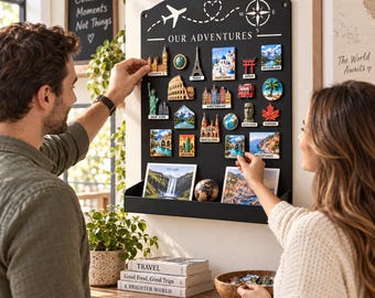 May include: A black metal wall-mounted display board with the text "OUR ADVENTURES" at the top, featuring a white airplane, heart, and compass graphic. The board holds various colorful travel-themed magnets and two rectangular photo prints.