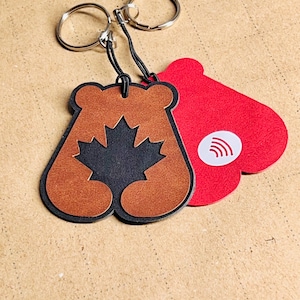 May include: Keyrings with silver rings. One keyring is brown with a black maple leaf design. The other keyring is red with a white circle and curved lines. Both keyrings are attached to a black cord.