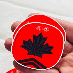 Può includere: Adesivi rotondi rossi e neri con un design a foglia d'acero nera. Gli adesivi hanno un bordo bianco e il testo "THIS STICKER IS NOT SPONSORED BY THE WHY NOT COMPANY". Gli adesivi sono tenuti in una mano.