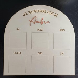 Peut inclure: Tableau de jalons en bois avec un dessus en forme d'arche. Le tableau comporte six espaces carrés et le texte "LES SIX PREMIERS MOIS DE Ambre". Le nom "Ambre" est en rose, en écriture cursive.