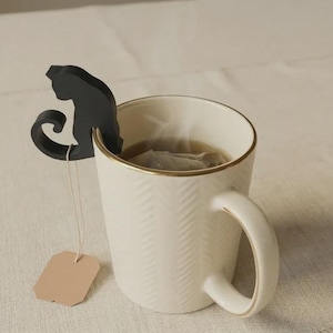 May include: A white ceramic mug with a gold trim, holding tea and a tea bag. A black cat-shaped tea infuser sits on the rim. A blank tag hangs from the tea bag. The mug has a textured pattern.