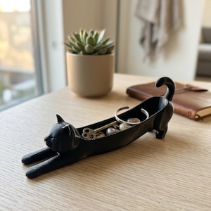 May include: A black cat-shaped tray with a hollowed-out back for storage. The cat is depicted in a stretching pose with its front paws extended forward and its tail curled upward. Inside the tray are a metal key and a silver-toned metal bracelet.