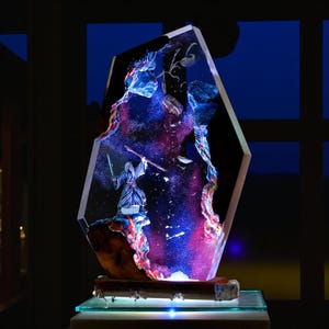 May include: An illuminated sculpture of a figure in a blue robe wielding a sword and staff, set against a cosmic backdrop of purple, blue, and red. The sculpture is encased in a clear, faceted form and mounted on a wooden base.