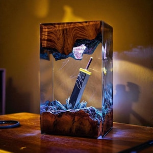 May include: A rectangular resin art piece featuring a black sword with a yellow hilt, set within a clear resin block. The base and top of the block have a wood grain design. Blue and green accents add detail to the piece.