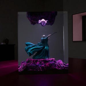 Può includere: Una scultura di un mago in una veste verde acqua, con una bacchetta, su una base strutturata viola e rosa. Una forma scura a forma di nuvola viola fluttua sopra. La scena è racchiusa in una struttura rettangolare trasparente.