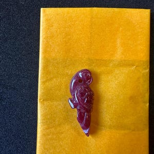 May include: A carved, deep red parrot pendant. The parrot is detailed with carved feathers and a curved beak. The pendant is set against a yellow background and a black surface.