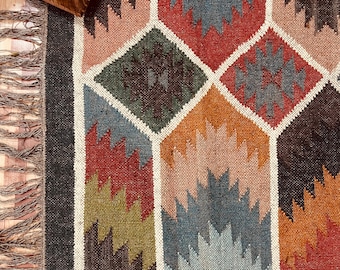 May include: A colorful geometric rug with a diamond pattern in shades of red, blue, orange, green, and brown. The rug has a fringed edge and a dark border. The design features a repeating pattern of zig-zag shapes within each diamond.