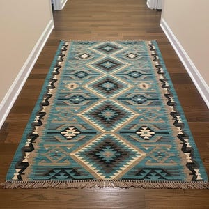 Pode incluir: Um tapete retangular turquesa com um padrão geométrico de losangos em tons de castanho, bege e preto. O tapete tem uma borda com franjas e está colocado sobre um chão de madeira. O design é simétrico e apresenta formas repetidas.