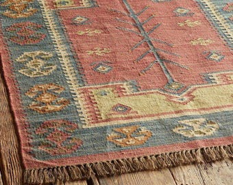 May include: A rectangular area rug featuring a central salmon-pink field with a dark blue geometric tree-like motif, surrounded by a cream border and a wide blue outer border with repeating orange and cream floral-inspired patterns and fringe.
