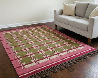 Puede incluir: Una alfombra rectangular con un patrón geométrico en verde oliva, crema y magenta. La alfombra tiene una textura tejida con un diseño de cuadrícula y largos flecos de color marrón oscuro en los bordes cortos, sobre un suelo de madera oscura.