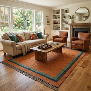 May include: A living room featuring a beige sofa with teal and orange pillows, two brown leather armchairs, and a wooden coffee table. A large orange area rug with teal borders and fringe sits on the hardwood floor. A fireplace and shelves are visible.