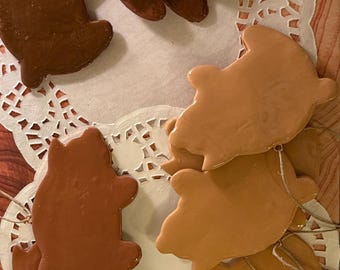 May include: A collection of bear-shaped ornaments in various shades of brown, from light tan to dark chocolate. Each ornament has a glossy finish and is strung with twine for hanging. They are arranged on a white doily.