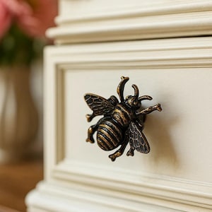May include: A decorative drawer pull in the shape of a bee, made of bronze-colored metal. The bee has detailed wings and a striped body. It is mounted on a cream-colored drawer front with a decorative frame.