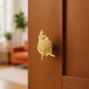 May include: A gold-colored seashell-shaped door knob is mounted on a brown wooden door. The knob has a detailed, textured surface, and the door has a vertical wood grain pattern. The background is blurred.