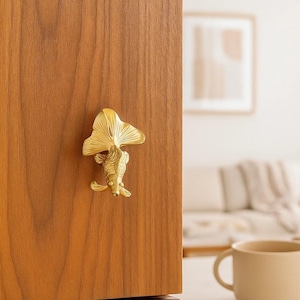 May include: A gold-colored fish-shaped cabinet knob mounted on a wooden cabinet door. The knob is a detailed sculpture of a goldfish with a fan-shaped tail. The wood grain of the cabinet is visible.