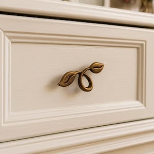 May include: Close-up of a cream-colored drawer with a decorative bronze-colored leaf-shaped handle. The drawer is part of a larger piece of furniture, possibly a dresser or vanity. The handle is the focal point.