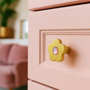 Może przedstawiać: Zbliżenie na różową szafkę ze złotą gałką w kształcie kwiatu. Gałka ma perłowy środek. Szafka ma zagłębiony panel. Różowa sofa widoczna w tle.