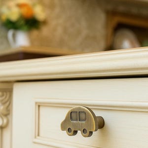 Puede incluir: Un tirador de armario de color bronce con forma de coche. El tirador está fijado a una puerta de armario de color crema. El diseño del coche presenta dos ventanas cuadradas y dos ruedas redondas. El fondo es una cocina borrosa.