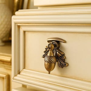 Op de afbeelding: Close-up van een crèmekleurige kastlade met een decoratieve messing eikelvormige trekker. De trekker heeft ingewikkelde details en is gemonteerd op een paneel ladefront met gouden rand. De kast heeft een klassiek, elegant ontwerp.