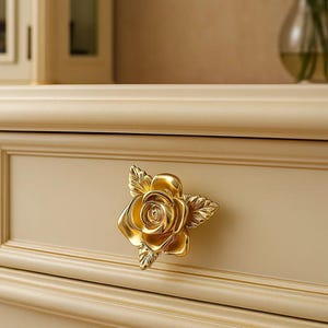 May include: A gold rose-shaped drawer pull on a cream-coloured cabinet. The cabinet features decorative moulding and a smooth surface. The rose has detailed petals and leaves, adding a touch of elegance to the furniture.