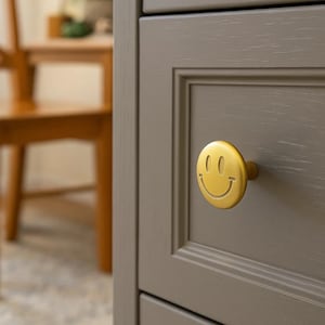 May include: Close-up of a gray cabinet drawer with a gold smiley face knob. The knob is round with a smiling face design. The cabinet has a recessed panel design.