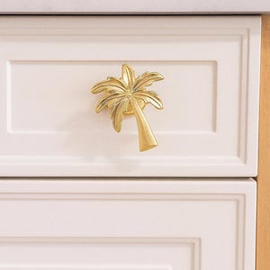 May include: A gold-colored palm tree cabinet pull is mounted on a white cabinet door. The palm tree design is detailed and three-dimensional. The cabinet door is part of a white kitchen cabinet.