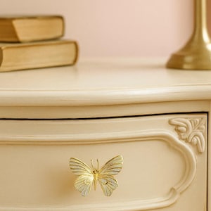 May include: A gold-colored butterfly-shaped drawer pull is centered on a cream-colored drawer. The butterfly has detailed wing patterns. The drawer is part of a cream-colored dresser with ornate detailing. A gold lamp and books are in the background.