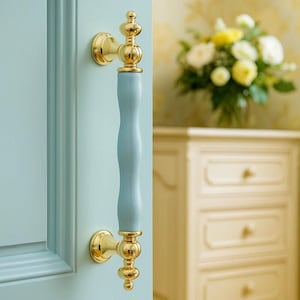 May include: A light blue door handle with gold detailing. The handle features a curved, pale blue central section and ornate gold ends. The door is light blue, and the background includes a white cabinet and flowers.
