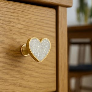 Könnte beinhalten: Ein herzförmiger Schrankknauf mit goldfarbenem Sockel und einem weißen, kristallartigen Zentrum. Der Knauf ist an einer hellen Holzschublade befestigt. Das Design verleiht Möbeln eine dekorative Note.