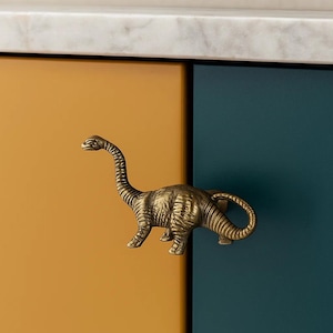 May include: A brass dinosaur-shaped cabinet pull is mounted on a mustard yellow and teal cabinet door. The dinosaur has a long neck and tail, with detailed texture.