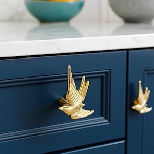 May include: Gold-colored bird-shaped cabinet hardware mounted on a navy blue cabinet. The bird is in flight, with detailed wing and tail feathers. The cabinet has a recessed panel design. A second bird-shaped handle is visible.