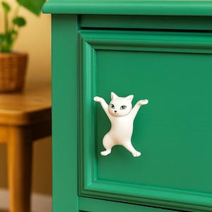 Peut inclure: Bouton de meuble blanc en forme de chat aux bras tendus, monté sur une porte d'armoire verte. Le chat a les yeux verts et est centré sur la porte. L'armoire est peinte en vert vif.