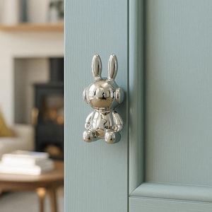 May include: A shiny silver bunny-shaped door knob is mounted on a light blue door. The door knob has a star on its chest and is in a sitting position. The background shows a glimpse of a living room.