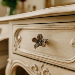 May include: Close-up of a cream-colored antique desk drawer with a bronze flower-shaped knob. The drawer features ornate carvings and a distressed finish, evoking a vintage aesthetic. A vase of red roses is slightly visible in the background.