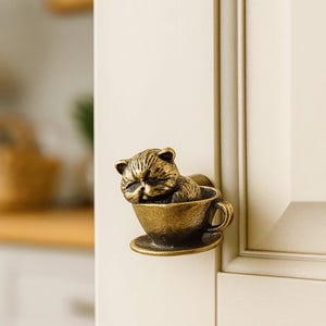 Peut inclure: Bouton de meuble en bronze représentant un chat endormi dans une tasse à thé. Le chat a des détails de fourrure et une expression détendue. Le bouton est monté sur une porte d'armoire crème.