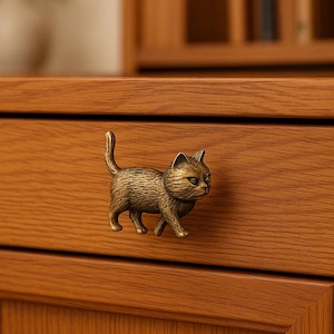 Puede incluir: Tirador de cajón con forma de gato de color bronce, montado en un cajón de madera. El gato está representado en una pose de caminar con la cola levantada. El cajón es de madera con veta visible.