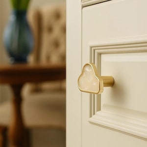 May include: A cloud-shaped cabinet knob with a gold finish and a pearlescent centre. The knob is mounted on a cream-coloured cabinet door. The background includes a blurred view of a vase with yellow flowers and a wooden table.