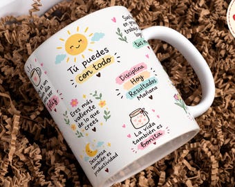 Puede incluir: Una taza de cerámica blanca con ilustraciones coloridas de un sol, una luna, flores y frascos, acompañada de frases motivacionales en español como "Tú puedes con todo", "Eres más valiente de lo que crees" y "La vida también es bonita."